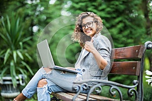 Happy brunette woman in eyeglasses sitting on the bench in park with laptop computer and showing thumb up