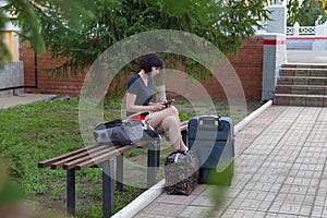 A happy brunette woman is sitting on a bench at the train station with a lot of bags suitcase waiting
