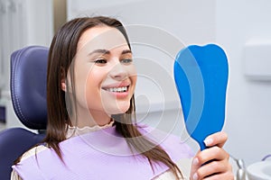 Happy brunette woman using mirror while looking at her teeth after dental treatment