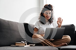 Happy brunette woman waving hand while taking video call on laptop