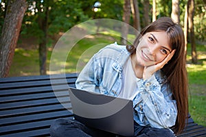 Happy brunette young girl freelancer sitting on bench in park with laptop