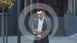 Happy businesman in sunglasses and a suit counting money and walking near office building. He selebrating his successful