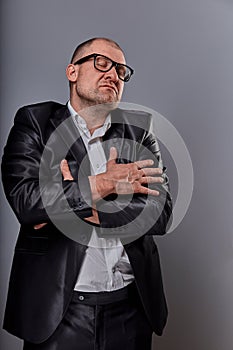 Happy business man in eye glasses hugging himself with natural emotional enjoying face with closed eyes on studio grey background