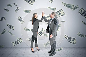 Happy business partners celebrating success standing under money rain of banknotes falling on wall background