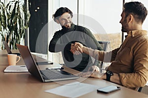 Happy business partners shaking hands in agreement, making deal, sitting at desk with laptop at modern office