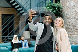 Happy business people, working in office. Smiling Caucasian lady and African man making joint selfie photo on smartphone