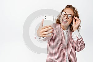 Happy business woman in glasses and suit taking selfie, laughing and smiling, using photo filter app on mobile phone