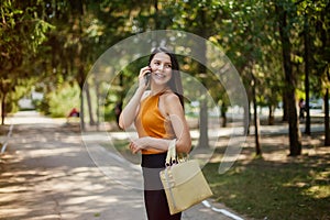 Happy business woman laughs, talking on the phone goes on the road in the park with a bag and looks back