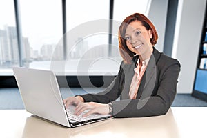 Happy business woman with red hair smiling at work typing on computer laptop at modern office desk