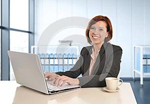 Happy business woman with red hair smiling at work typing on computer laptop at modern office desk