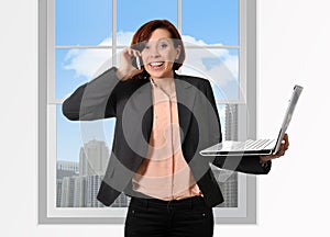 Happy business woman with red hair talking on the mobile cell phone holding computer laptop in hand multitasking