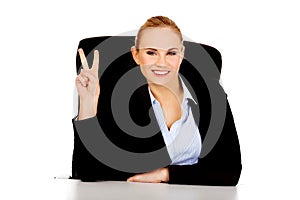 Happy business woman sitting behind the desk and shows victory sign