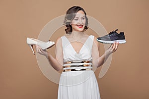 Happy business woman in white dress and holding sneakers and heel and toothy smiling at sporty sneakers. choosing comfortable