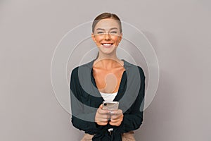 Happy business young woman posing isolated over grey wall background using mobile phone.