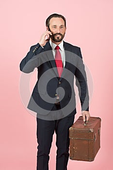 Businessman in black suit and red tie with vintage briefcase talking over phone on pink wall background.