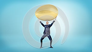 A happy businessman on blue background holds a huge golden egg over his head.