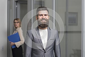 Happy businessman with blurred woman on background. Bearded man in formal suit in office. Confident man smile with beard