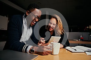 Happy businessman and businesswoman sitting together laughing while looking at digital tablet during break time at night