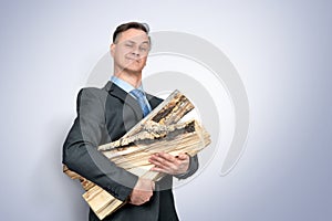 Happy businessman in a dark suit and tie holds firewood in his hands on light blue background. Heating concept.