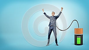 A happy businessman with raised hands is connected to a large battery charging him.
