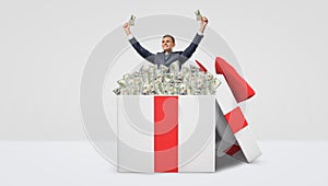 A happy businessman standing inside a large gift box full of dollar bills with his hands raised in victory motion.