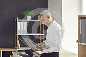 Happy businessman standing by office desk, holding laptop and doing some work online