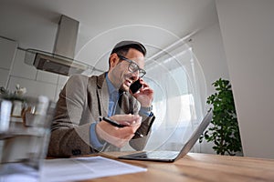 Happy businessman in suit having business call on smart phone and working over laptop at home office