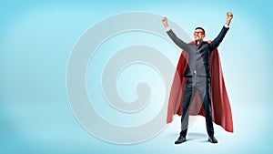 A happy businessman in a superhero red cape standing in victory pose on blue background.