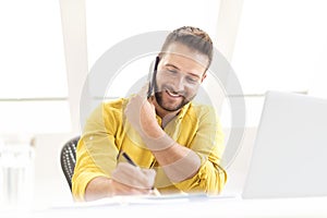 Happy businessman talking with somebody on his phone and making a notes while working in the office