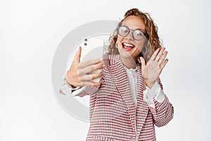 Happy businesswoman in glasses having video call conversation, waving at mobile phone screen and smiling friendly, white