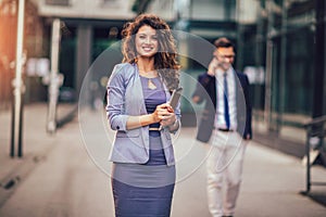 Happy businesswoman holding digital tablet outside of modern building, businessman in background