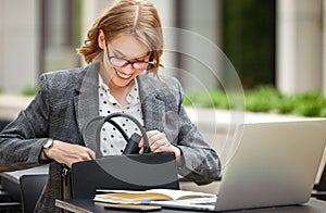 Happy businesswoman looking for something in handbag while working outside with laptop