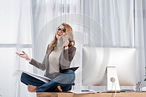happy businesswoman with papers talking on smartphone while sitting on table