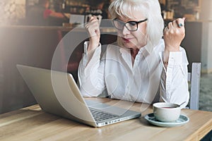 Happy businesswoman sitting in cafe at table in front of laptop,holding hands up and looking at monitor.Good news.