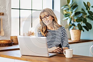 Happy businesswoman sitting in the kitchen and using laptop while having video conference