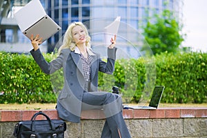 A happy businesswoman on the street holding a laptop and office paper. A woman sits curbside.