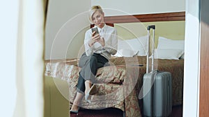 Happy businesswoman talking with family via online video chat using smrtphone camera sitting on bed in hotel room