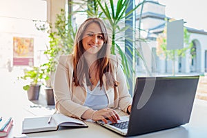 Happy businesswoman working online in office using laptop. Smiling middle-aged woman looking at camera sitting at desk
