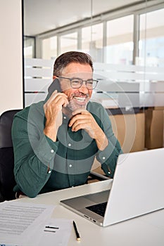 Happy busy mature business man making phone call using laptop in office.