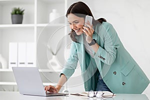 Happy busy millennial caucasian lady in suit typing on computer, calls by phone at workplace