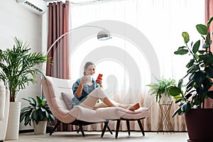 Happy calm Caucasian woman relax sit in comfort chair in cozy home interior use smartphone. Smiling young woman use