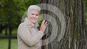 Happy calm mature middle aged single woman looking at camera posing near large tree, smiling older female, healthy