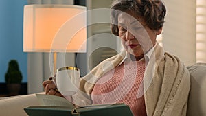 Happy calm peacefully Caucasian old woman enjoying literature in living room covered with blanket on sofa drinking tea