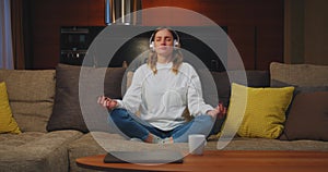 Happy calm young woman in headphones listen to relaxing music for yoga meditation, sitting in lotus pose on sofa at home