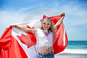 Happy Canadian girl carries fluttering white red flag of Canada against blue sky and ocean background.