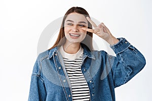 Happy candid girl, smiling and looking aside at promotional text, reading copyspace while showing peace v-sign, standing