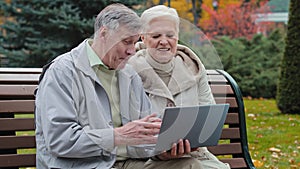 Happy carefree elderly senior grandparents sit on bench in autumn park man typing on laptop together reading discussing