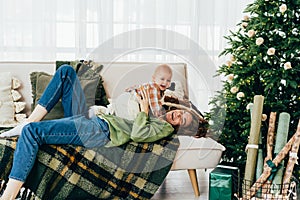 Happy carefree mom and baby are lying on the couch next to the Christmas tree and laughing