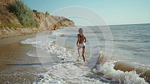 happy carefree slender woman with beautiful fitbody running along seashore of sea with waves after storm, girl jogging