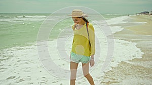 Happy carefree woman in hat walks alons sea water at sandy beach, enjoying find, sea waves and cloudy sky background. go